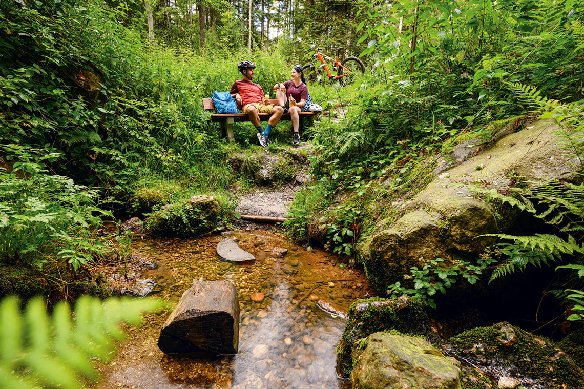 Radler-Paar rastet auf einer Bank am Bach im Wald bei Bad Birnbach, E-Bike im Hintergrund