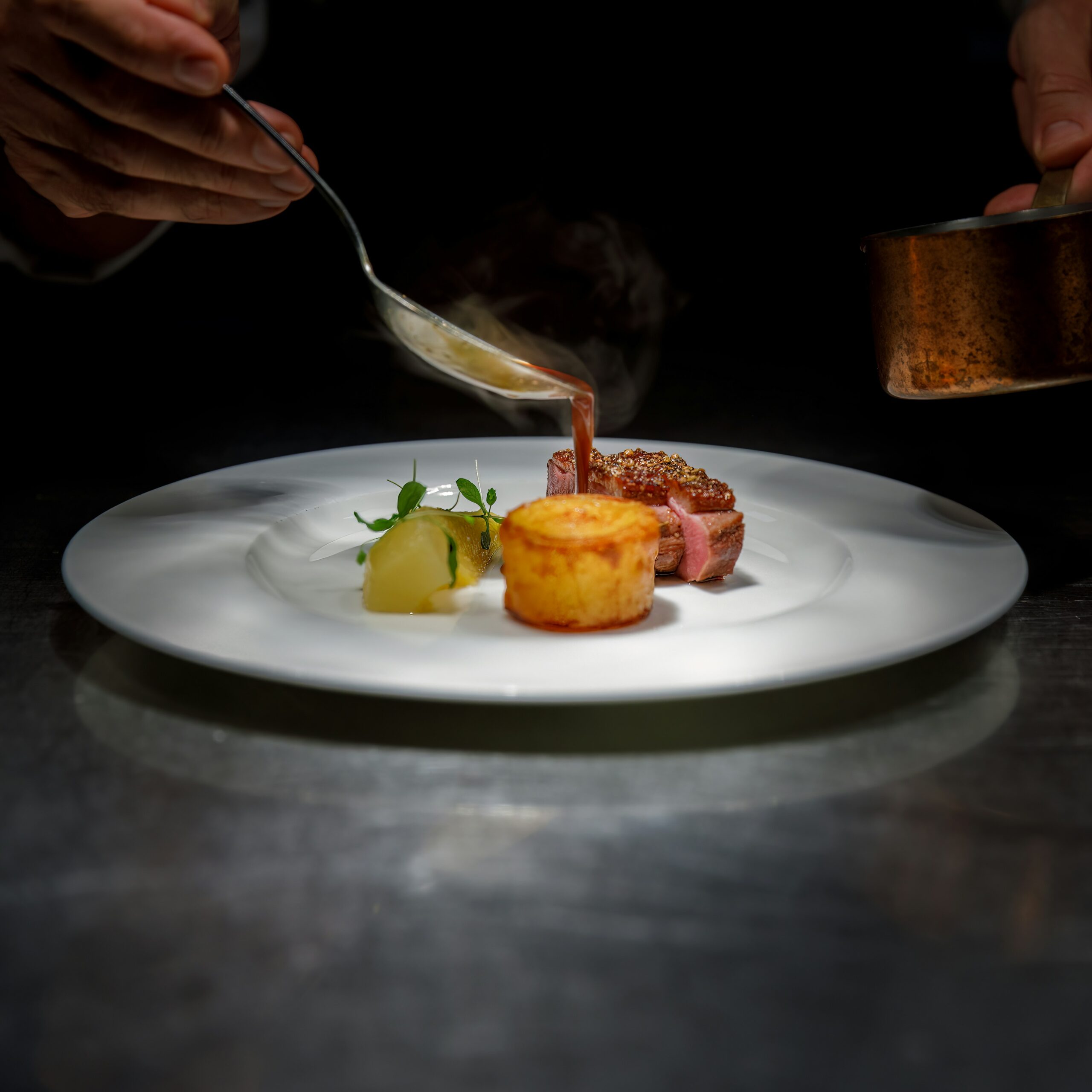Koch richtet ein Gourmetgericht an und gießt Sauce über zartrosa gebratenes Fleisch auf weißem Teller