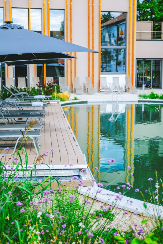 Naturpool mit Holzdeck, Liegen und Sonnenschirm vor der Fassade des Hotel Churfürstenhof
