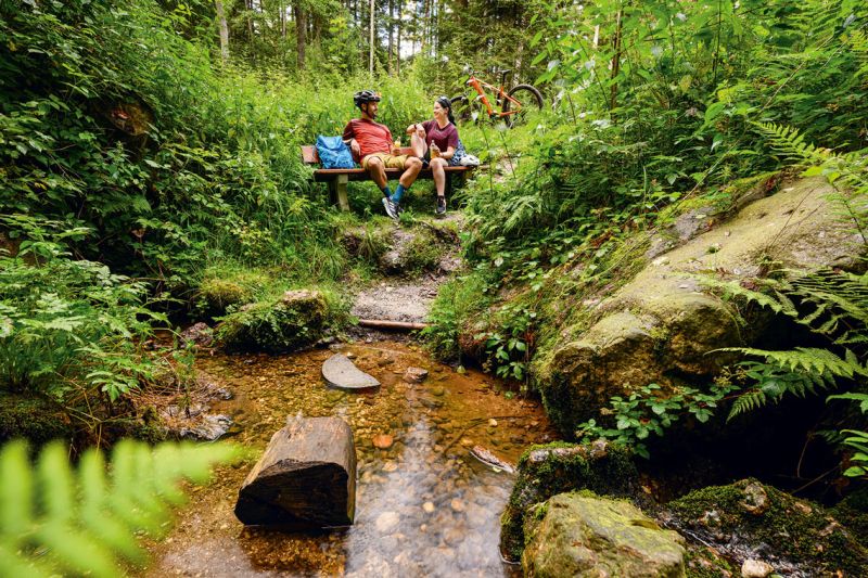 Radler-Paar rastet auf einer Bank am Bach im Grafenwald bei Bad Birnbach, E-Bike im Hintergrund