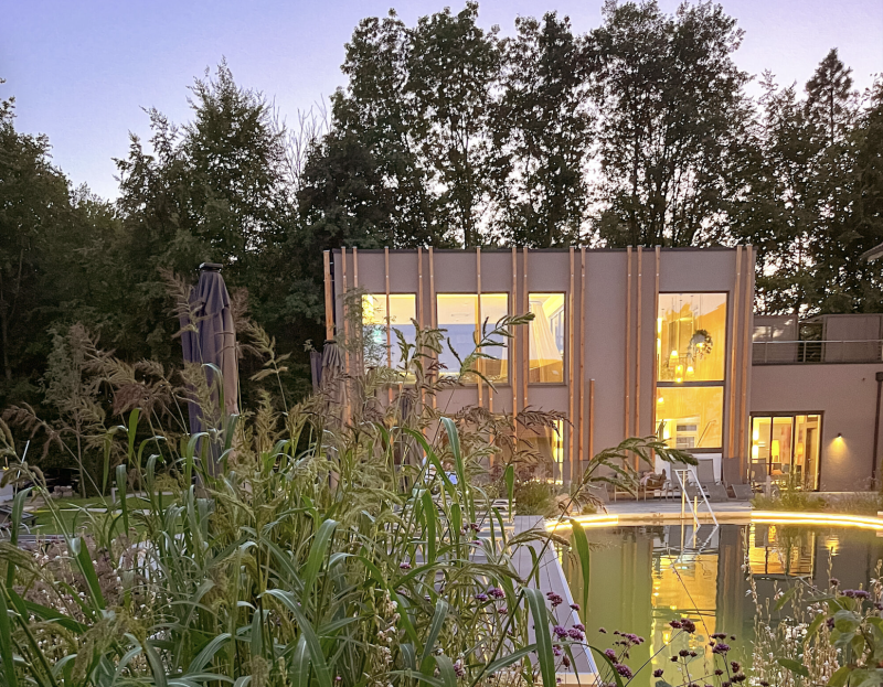 Stimmungsvoll beleuchtetes Badehaus mit Naturpool und Gräsern in der Abenddämmerung