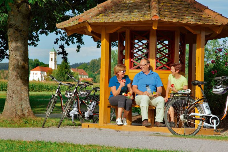 Radfahrer rasten in einem Holzpavillon bei Bad Birnbach, im Hintergrund eine Kirche